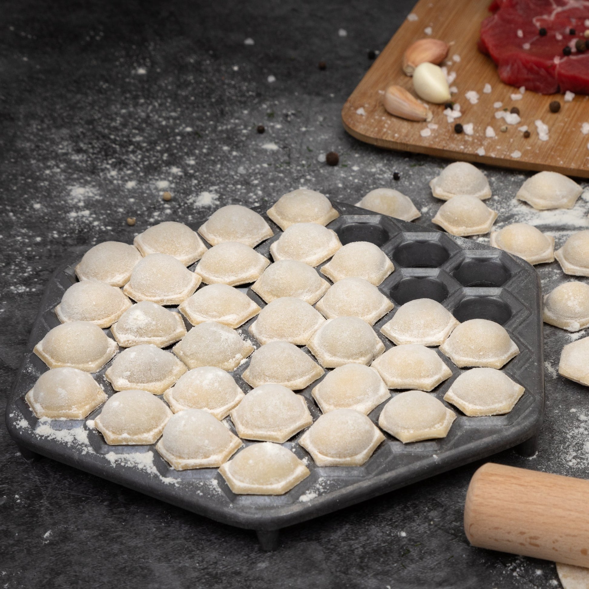 Uncooked pelmeni dumplings on mold with beef, garlic, and spices—Russian dumpling prep on dark surface.