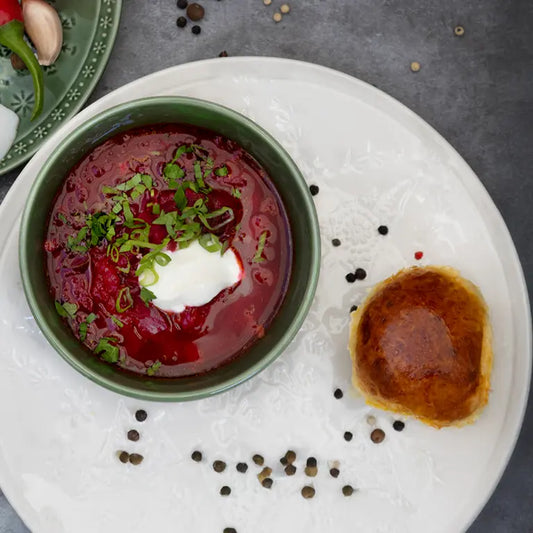 A top-down view of a bowl of red borscht soup garnished with chopped green onions and sour cream, served on a white plate next to a small golden-brown pastry, with scattered peppercorns on a gray surface.